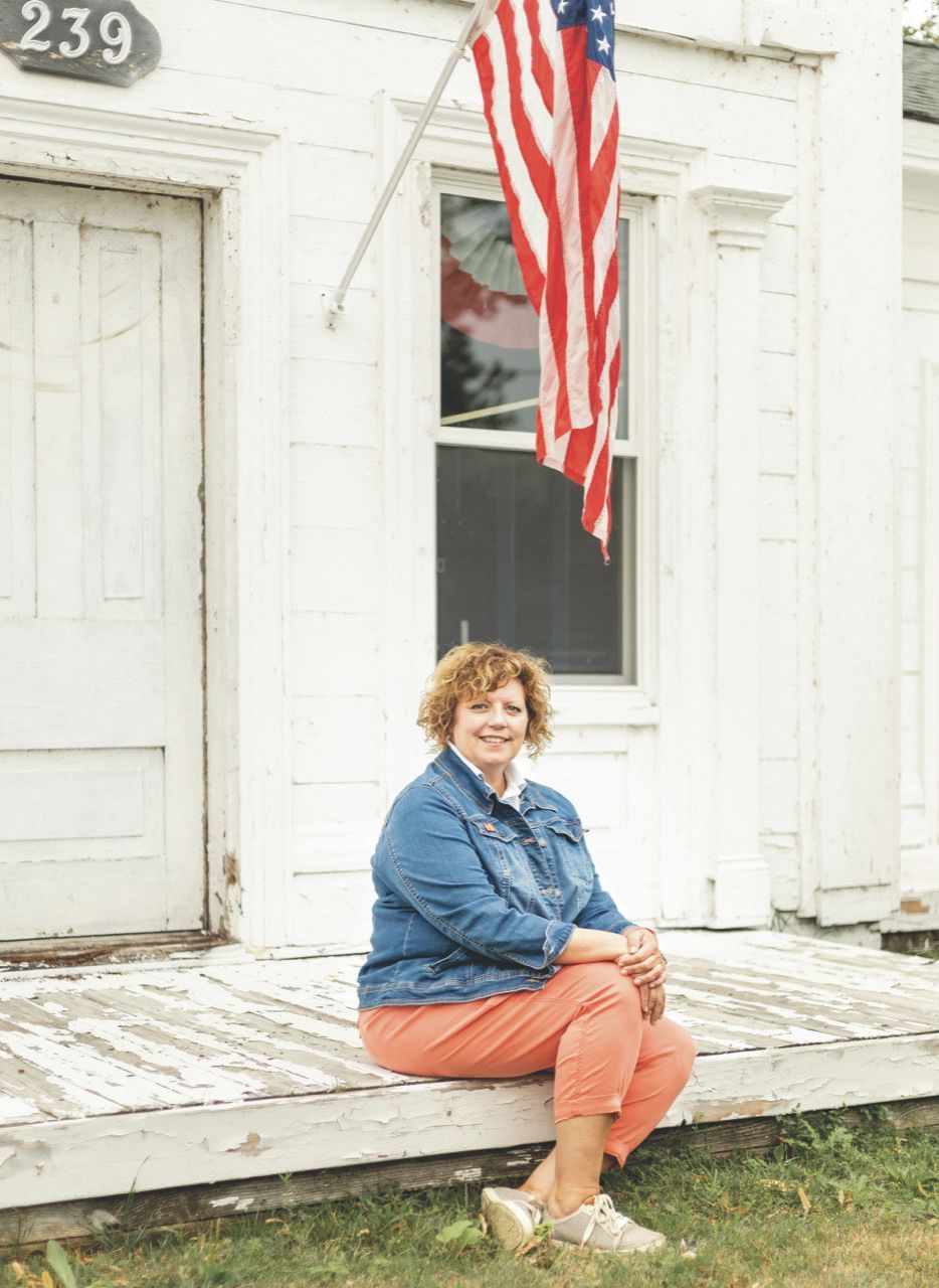 Mindy Fernandessitting on the porch of a white house with an American flag. She wears a denim jacket and orange pants.