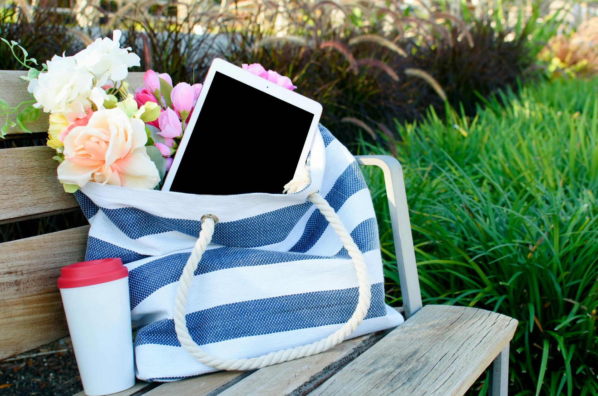A striped bag with a tablet and flowers in it is sitting on a wooden bench.