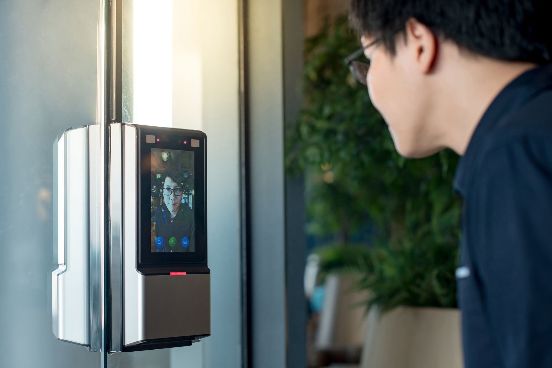 Man facing a facial recognition security device on a glass door. Device screen displays a person.