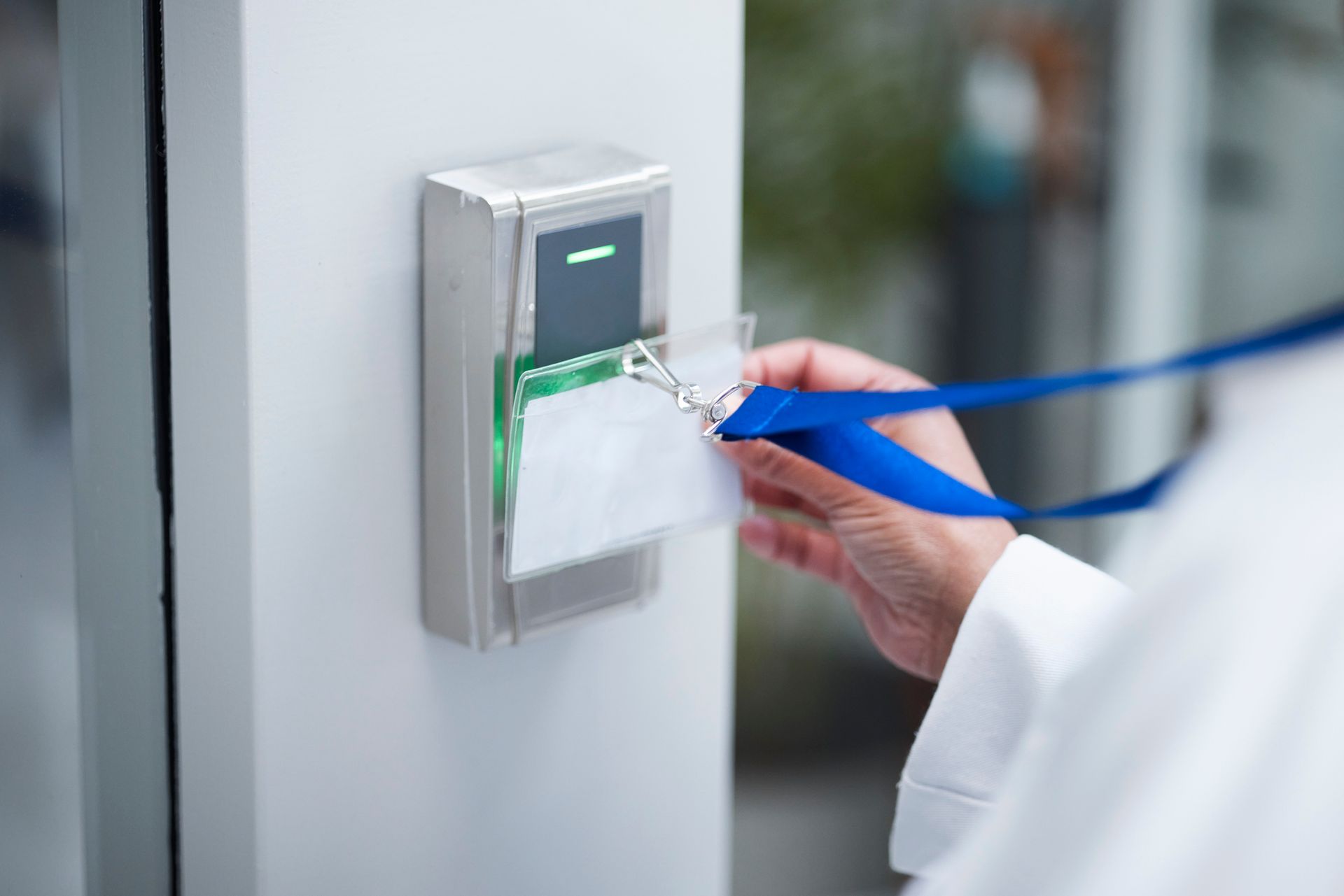 Person swipes a white ID badge near a door access reader, green light glows, blue lanyard.