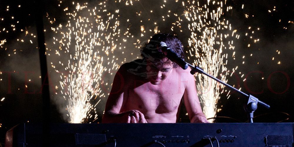 Un uomo a torso nudo canta in un microfono su un palco con fuochi d'artificio sullo sfondo.