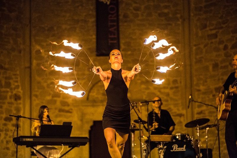 Una donna con un abito nero sta facendo giochi di prestigio con dei bastoncini di fuoco su un palco, con una band dietro di lei.
