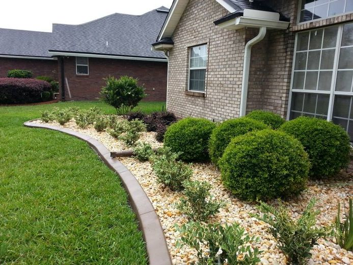 A manicured garden bed with rounded shrubs and light gravel, bordered by a concrete edge against a brick house.