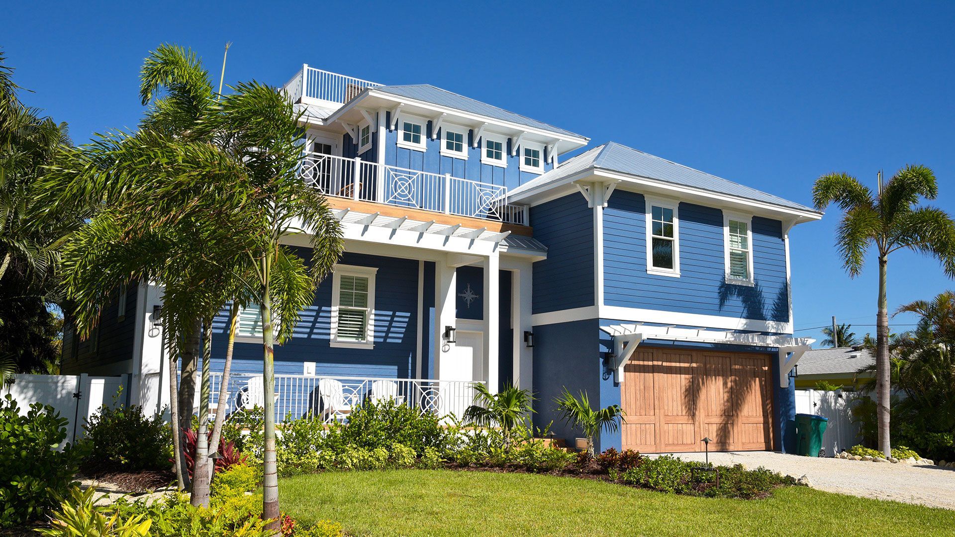 Blue two-story suburban house with white trim, garage, palm trees, and a sunny front yard