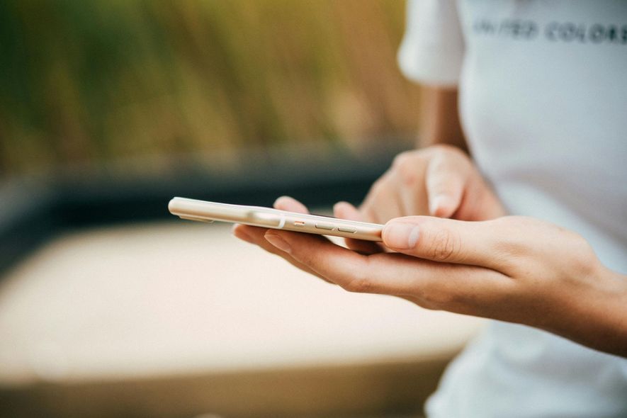 A close-up of hands holding and using a light-colored smartphone outdoors with a blurred background.