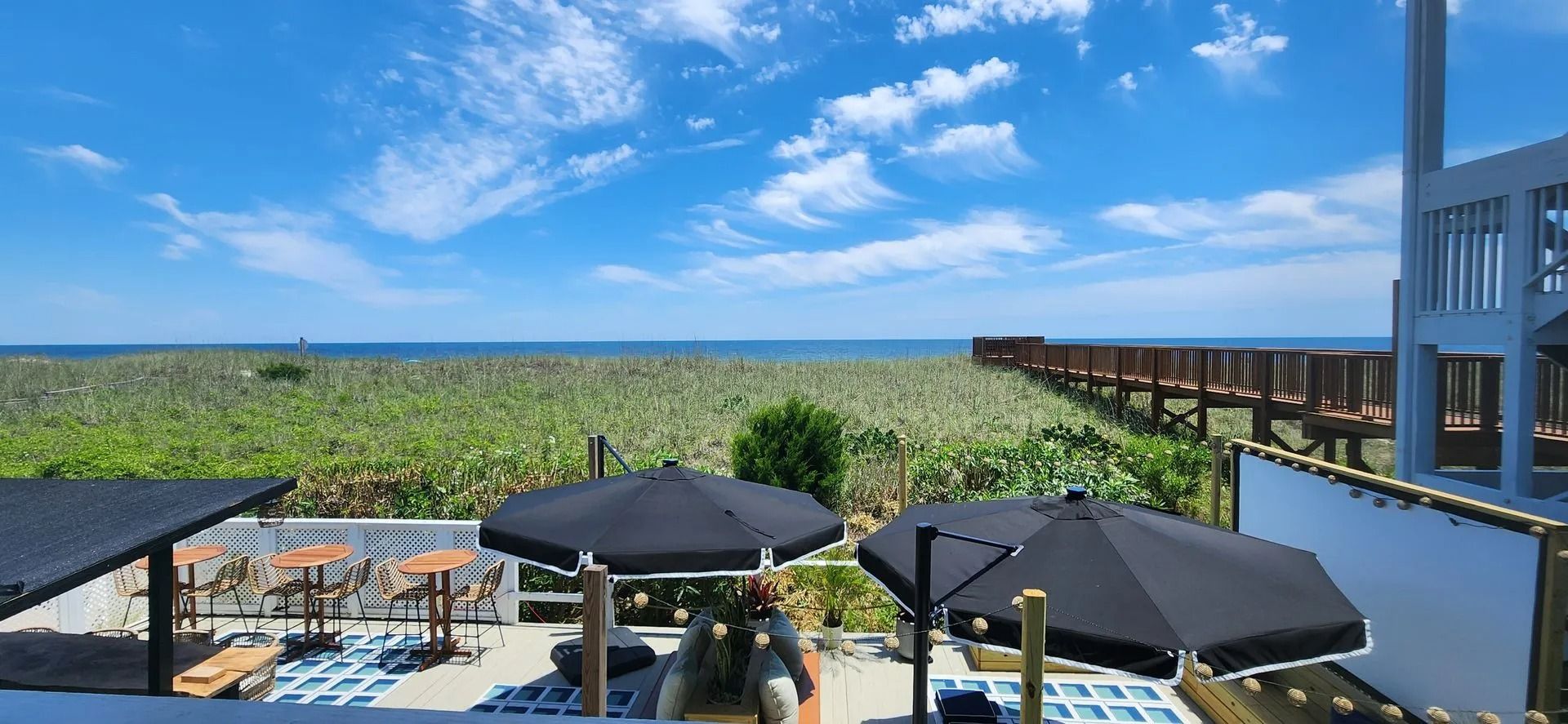 A patio with black umbrellas overlooks a sandy beach, green dunes, a wooden walkway, and the blue ocean under a sunny sky.