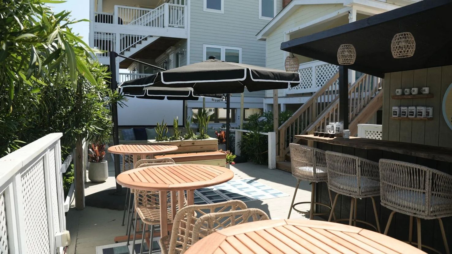 An outdoor patio with round wooden tables, wicker chairs, and black umbrellas, set beside a multi-story building.