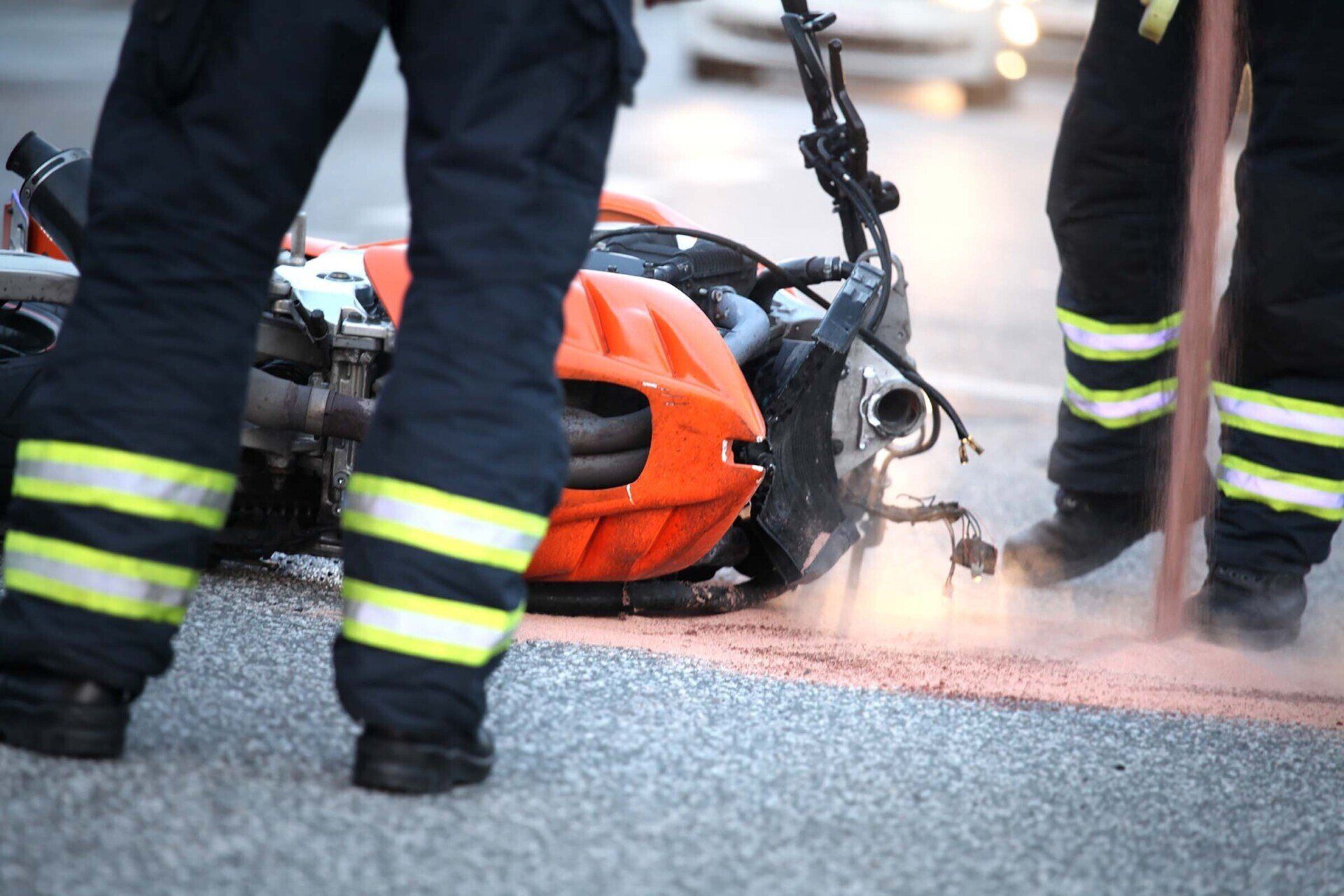 Firefighters at the scene of a motorcycle accident, orange bike on the ground, reflective stripes on pants.