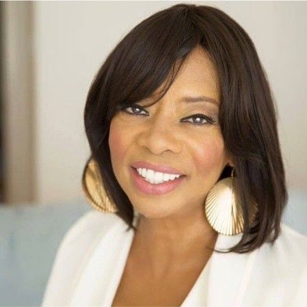 Singer, Helen Baylor wearing a white jacket and gold earrings is smiling for the camera.