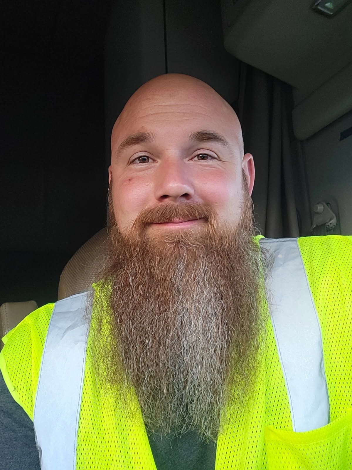 Bald man with long red beard, wearing a reflective safety vest, inside a truck cab.