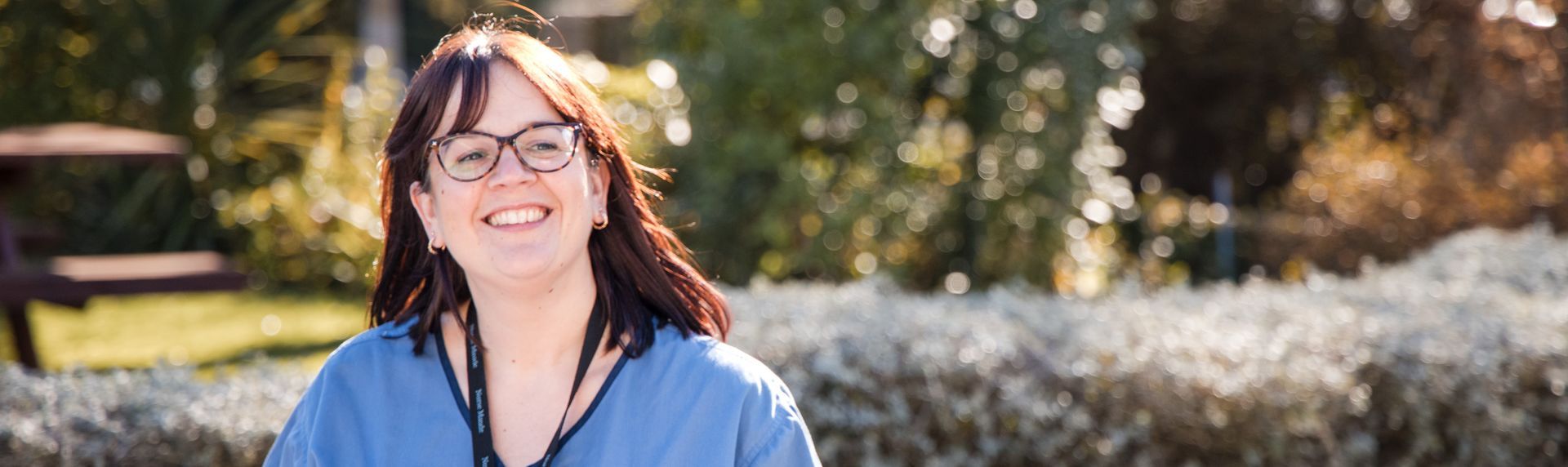 A woman wearing glasses and a blue scrub top is smiling for the camera