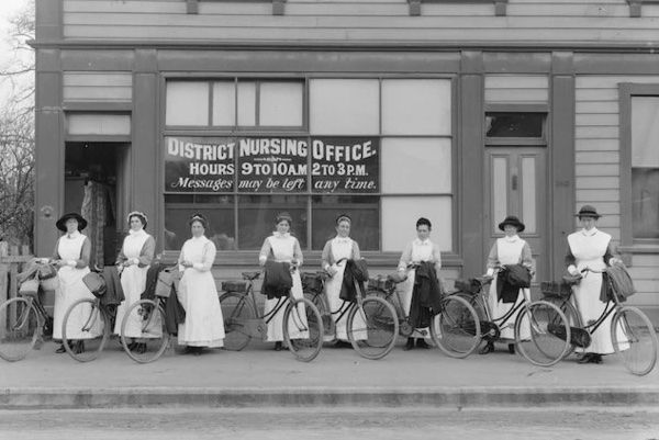 A group of women are standing in front of a building holding bicycles. Nurse Maude