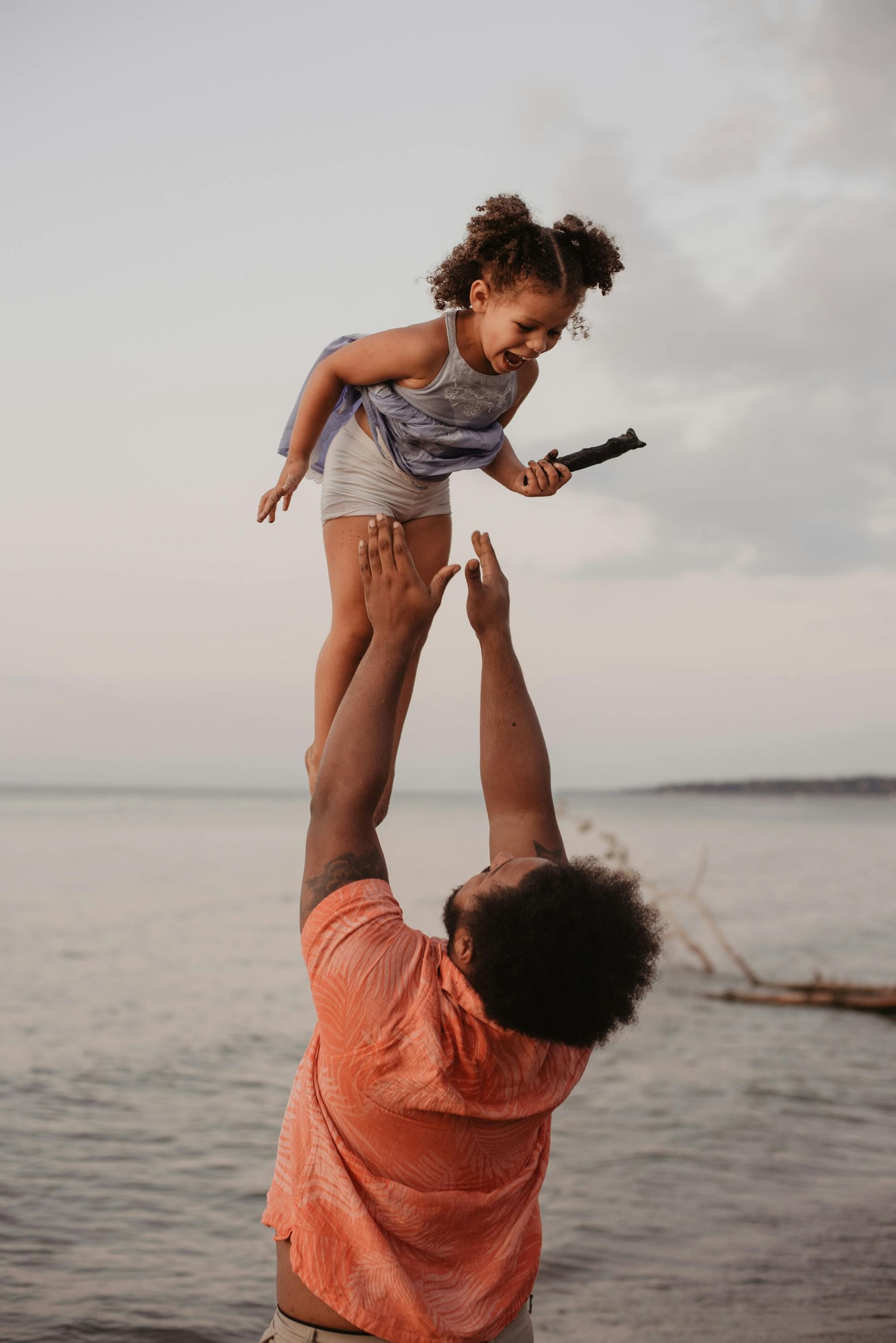 A man is holding a little girl in the air on the beach.