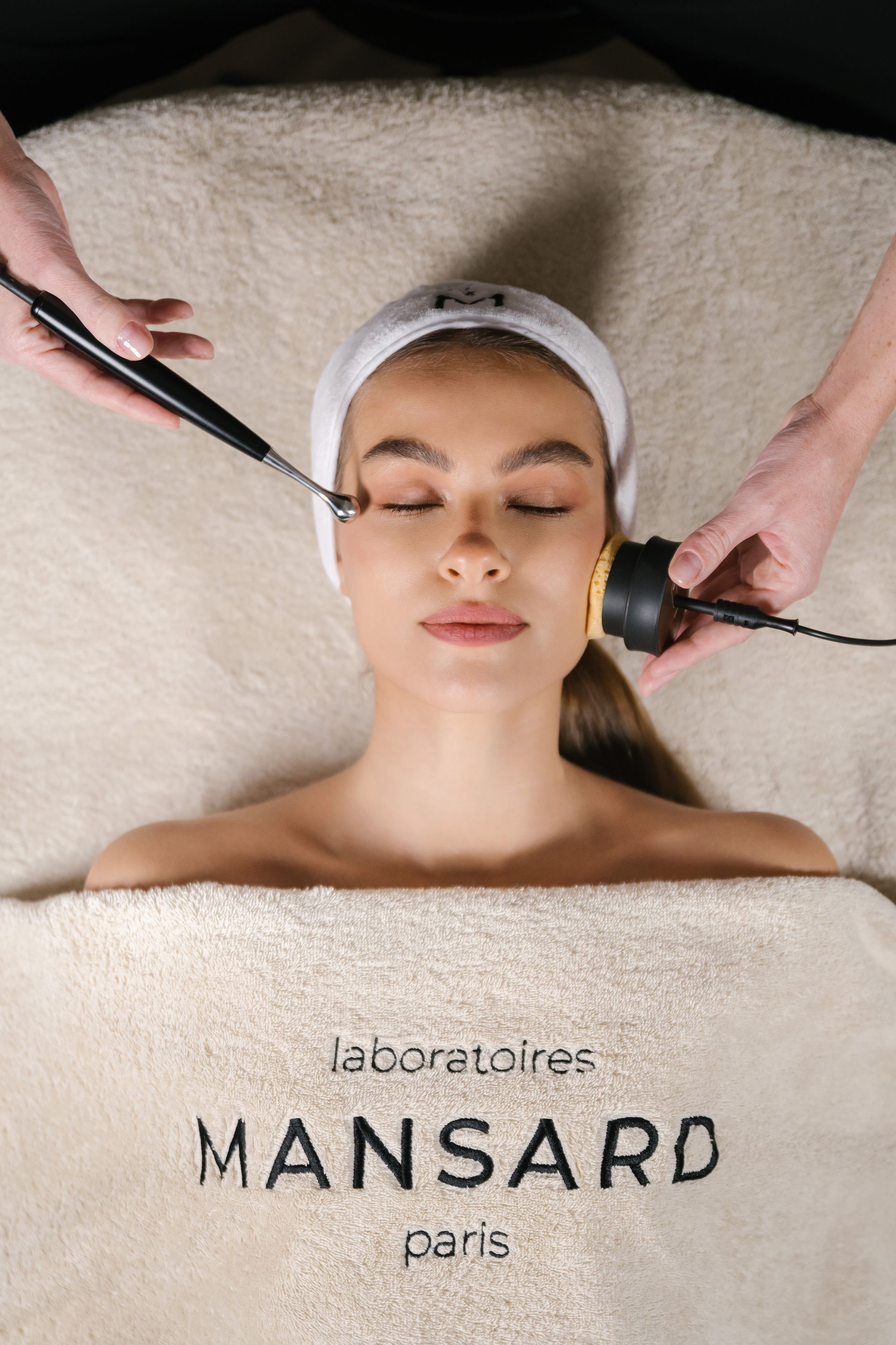 Une femme reçoit un soin du visage, allongée sur une table de spa, avec deux appareils tenus près de son visage.