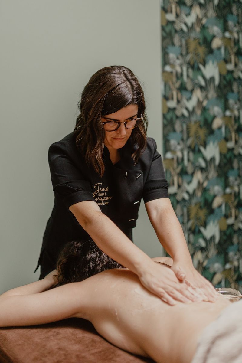 Une femme donne un massage du dos. Elle porte des lunettes et un haut noir. Les murs de la pièce sont vert clair.