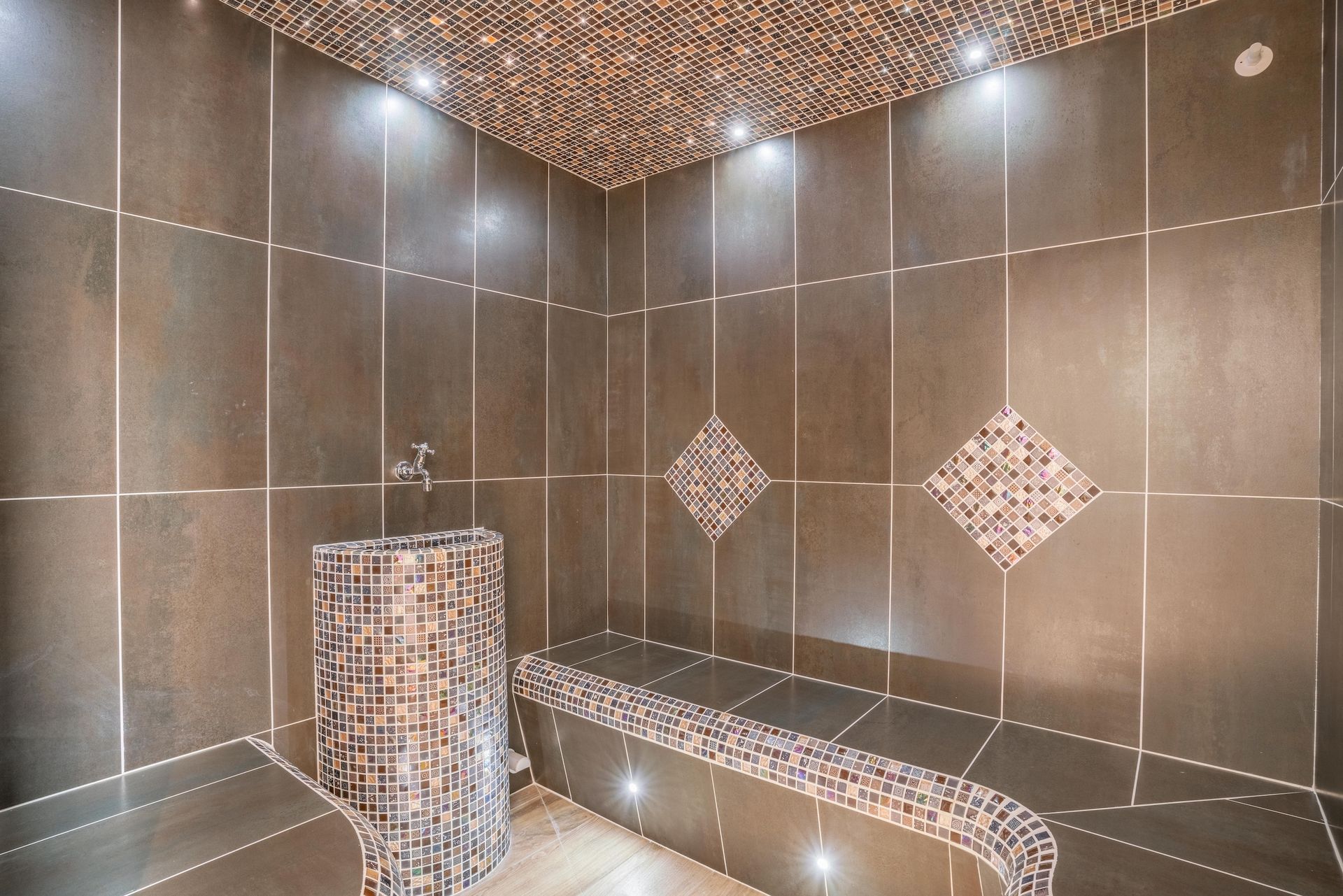 Hammam carrelé de faïence foncée avec accents de mosaïque, éclairage encastré et banquette.