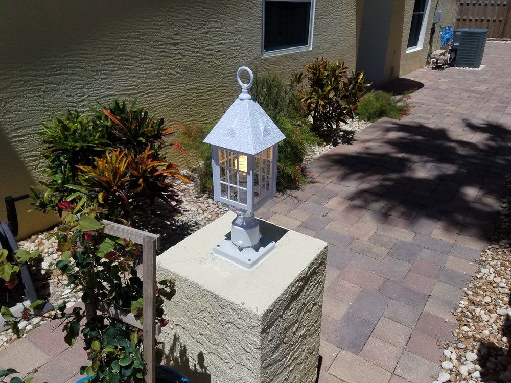 A white lantern lamp sits on a beige stone pillar along a paver walkway near a building exterior and garden plants.