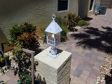 A white lantern lamp sits on a beige stone pillar along a paver walkway near a building exterior and garden plants.