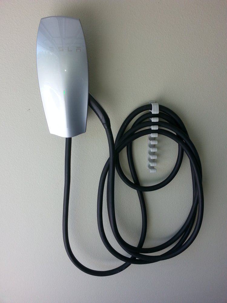 A silver Tesla Wall Connector electric vehicle charger mounted on a plain wall with a black coiled charging cable.