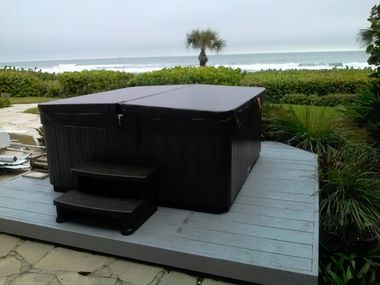 A dark brown hot tub with a cover sits on a gray wooden deck overlooking the ocean with a single palm tree in the distance.