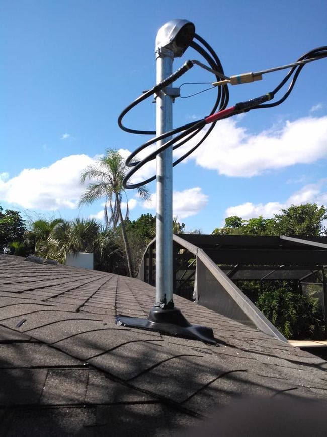 A metal utility mast with thick black electrical cables attached to a residential roof against a clear blue sky.