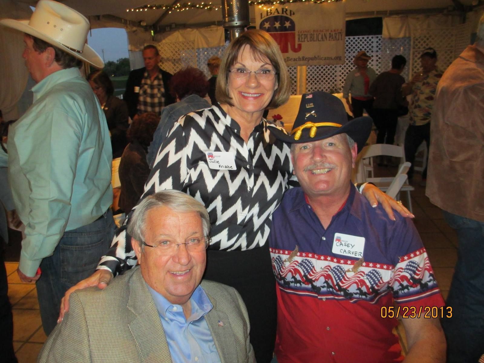 People seated at tables inside a tent, socializing and eating. Some have hats; American flags on the tables.