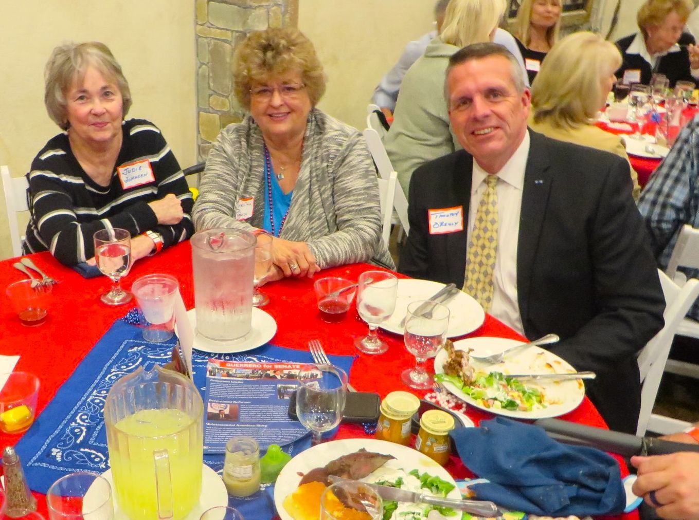 Three people at a table with food and drinks. The table is red.  They have name tags and are smiling.
