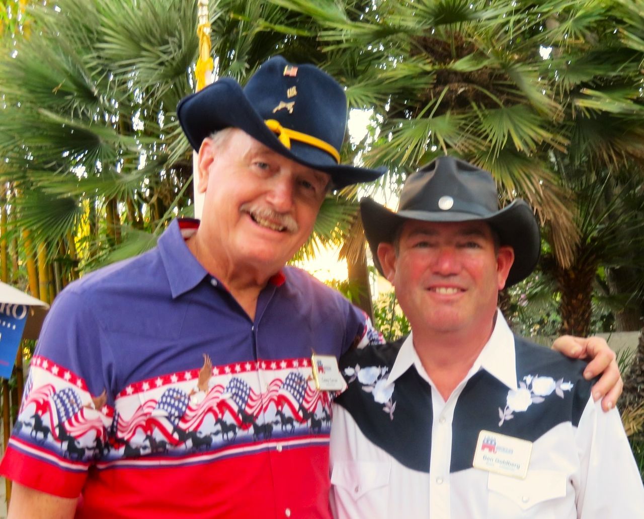 Two men in cowboy hats and patriotic shirts smiling outdoors, arms around each other.