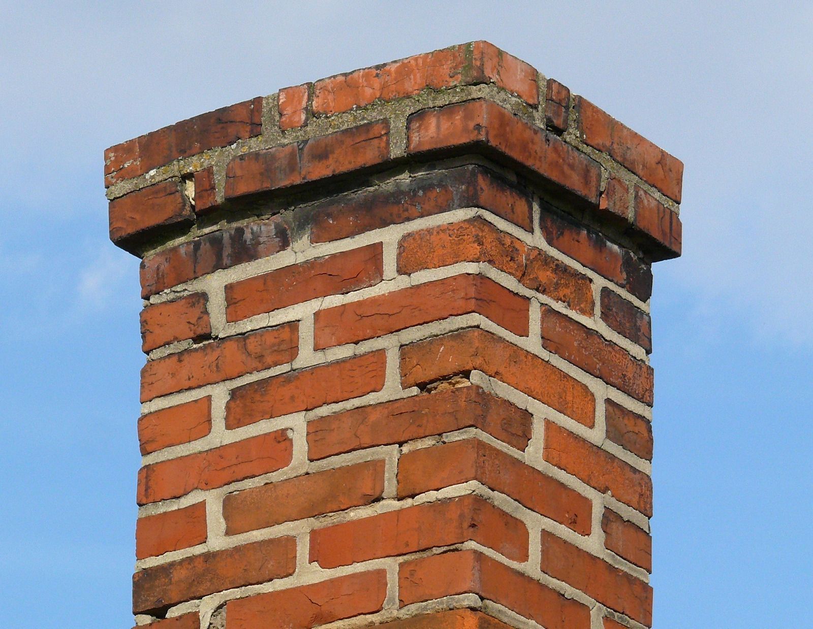 Brick chimney against a blue sky; weathered red bricks, dark staining near the top, square shape.
