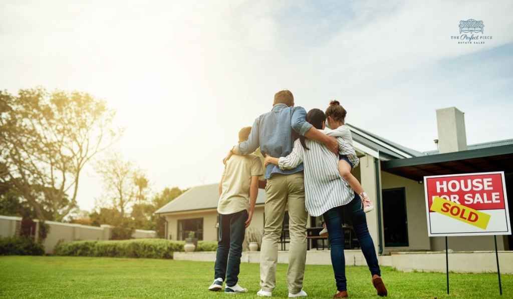 A family is hugging in front of a house for sale sign.