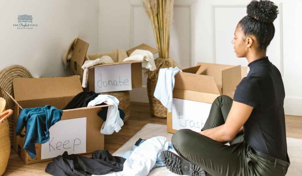 A woman is sitting on the floor in front of a pile of cardboard boxes filled with clothes.