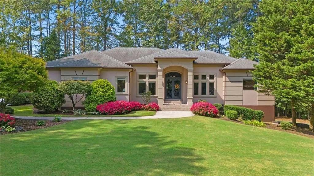 Single-story beige house with gray roof, green lawn, and pink flowering bushes.