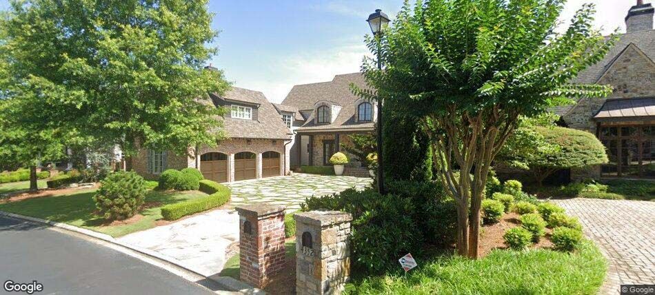 A large stone house with a driveway lined with trees and bushes.