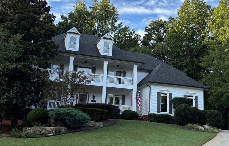 White two-story house with black roof, columns, and front balconies, surrounded by green trees and grass.