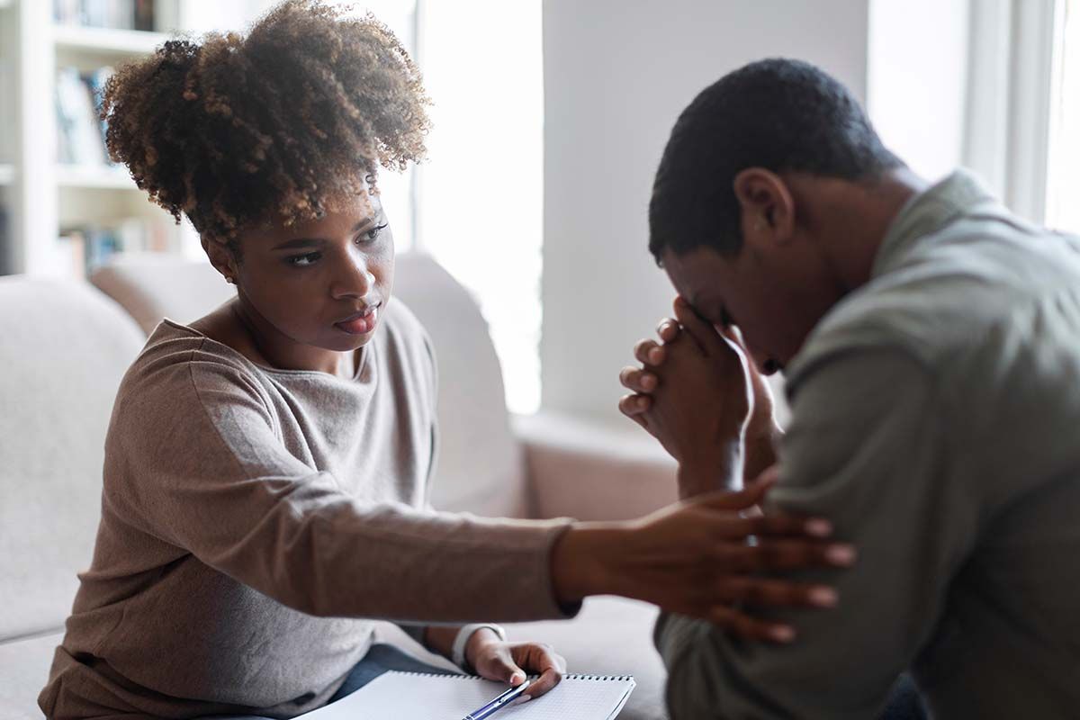 Woman comforting a person with hands clasped, perhaps in therapy. They are seated indoors.
