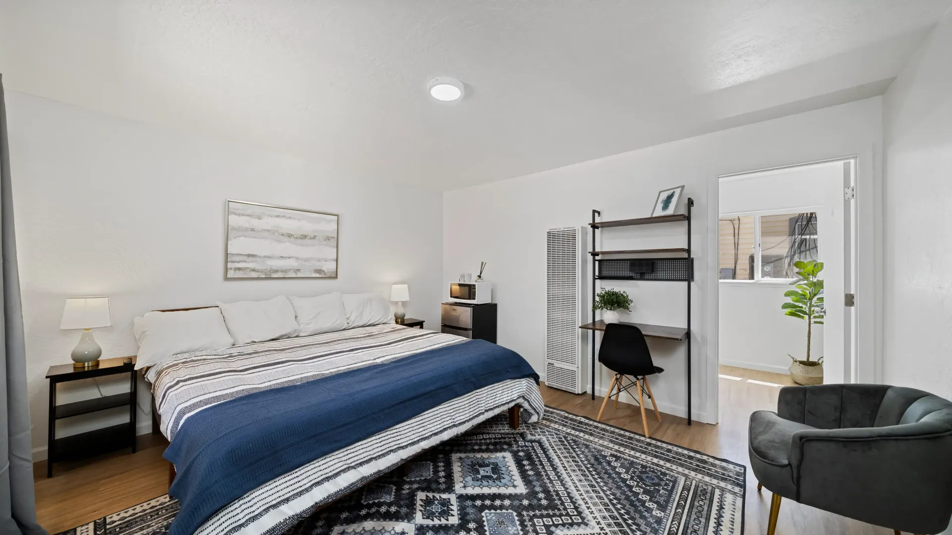 Bedroom with a bed, rug, desk, and armchair. White walls, blue and black accents, bright and airy.