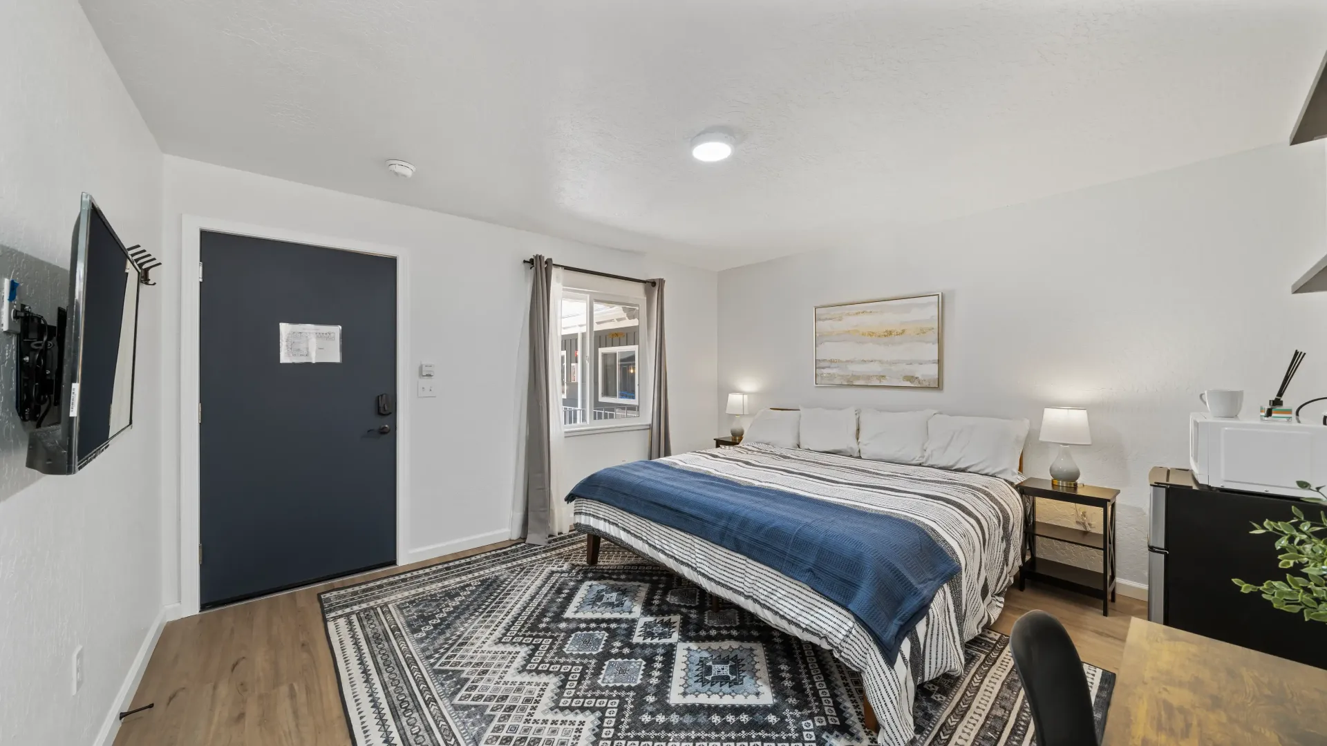 Bedroom with a bed, patterned rug, and dark blue door. White walls and wood flooring.