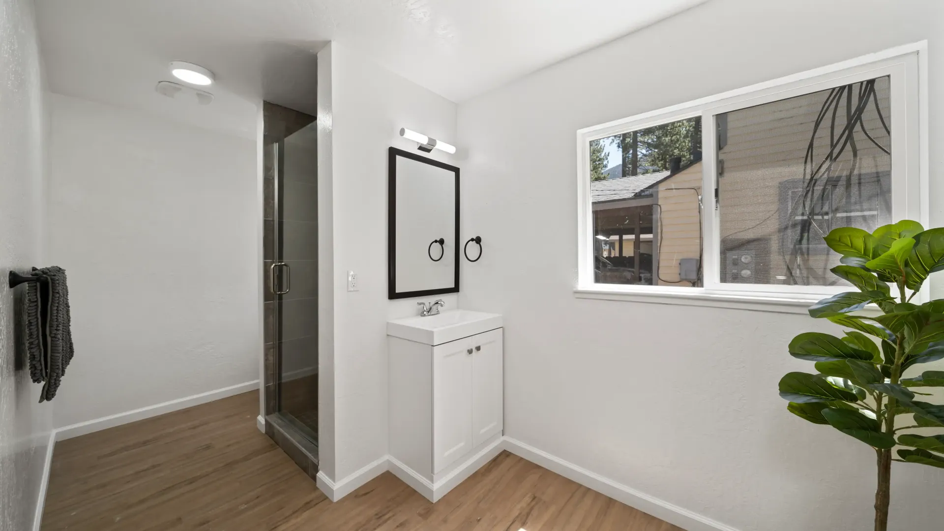 White bathroom with a window, a sink, and a shower. There's a plant in the corner.
