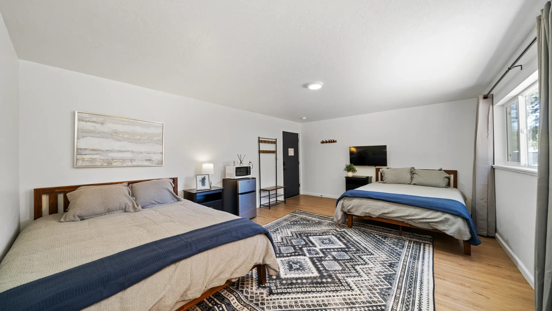 Two beds in a white-walled room with wood floors, a patterned rug, and a window with curtains.