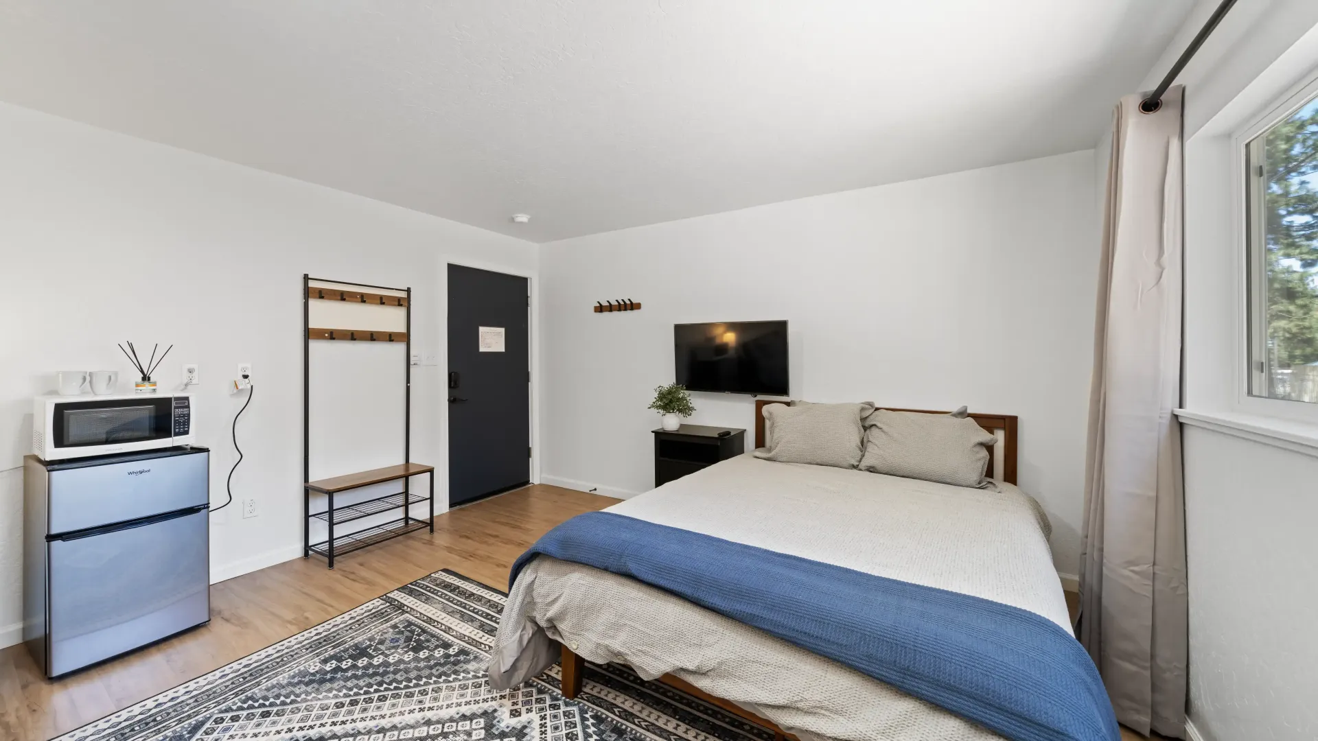 Bedroom with bed, TV, mini fridge, and a window. The room has white walls and a black door.