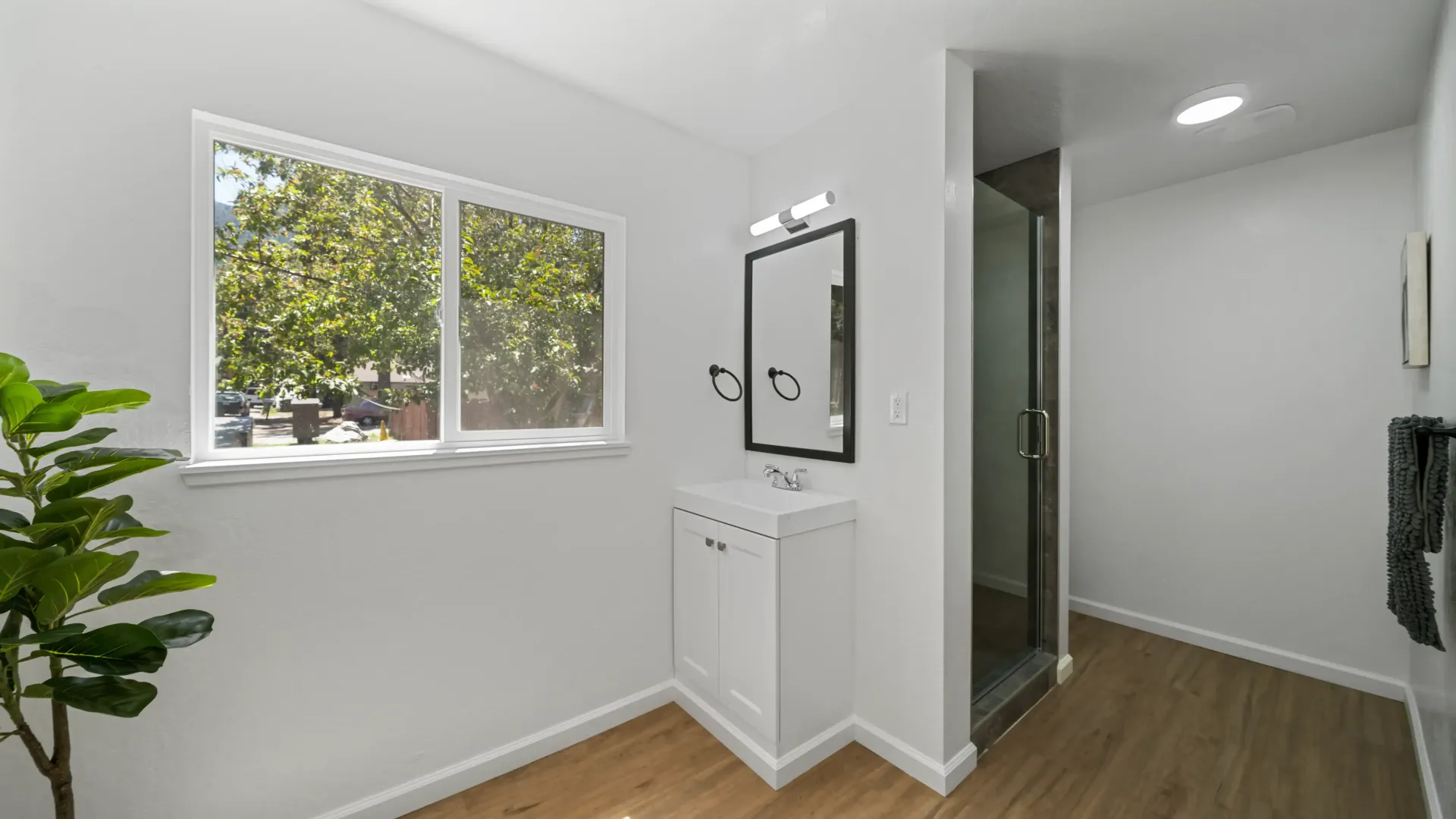 Bathroom with a window, white walls, wooden floors, white vanity, a mirror, and a glass shower.