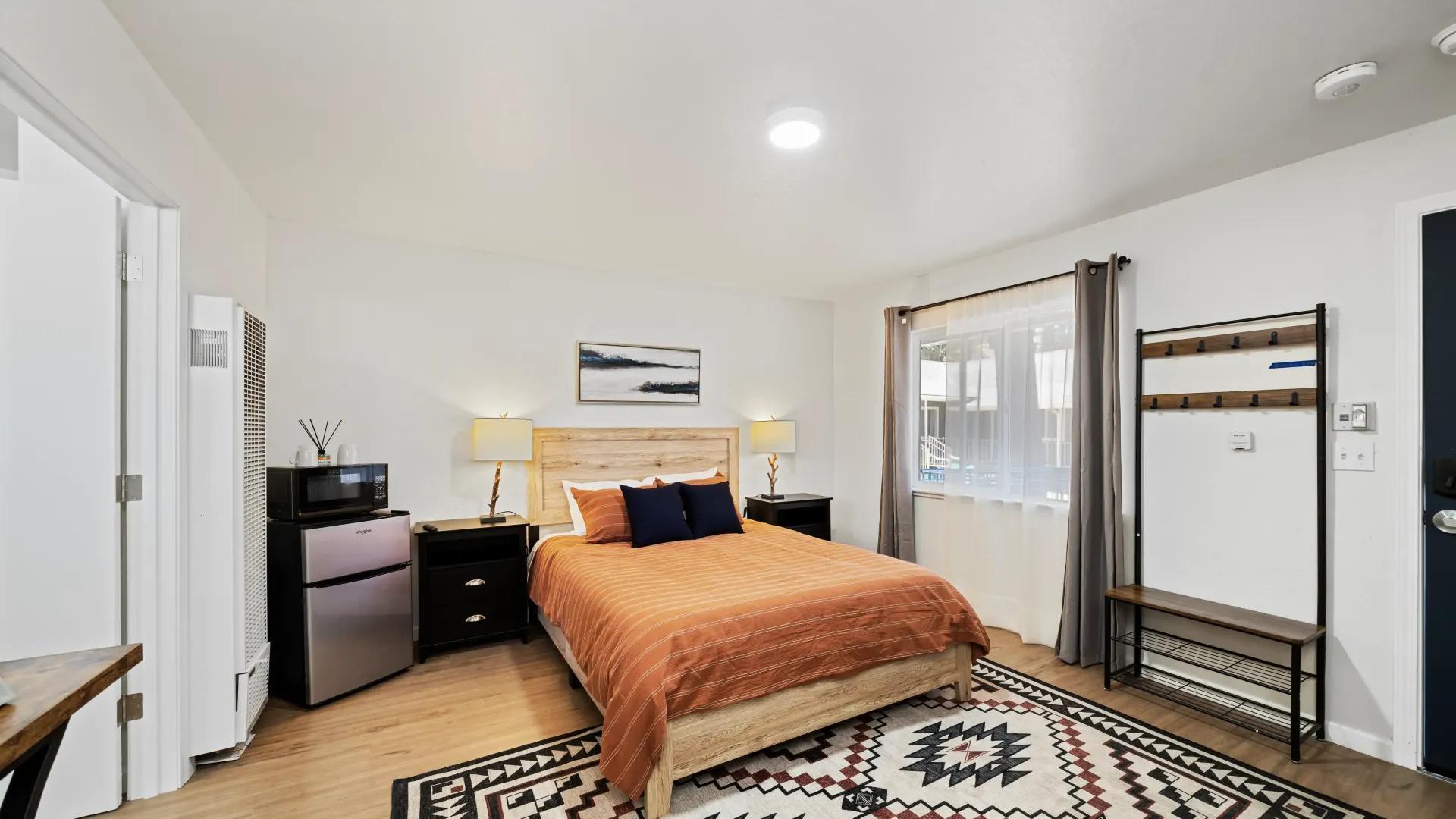 Bedroom with bed, nightstands, rug, mini-fridge, and clothes rack. White walls, neutral colors, and natural light.