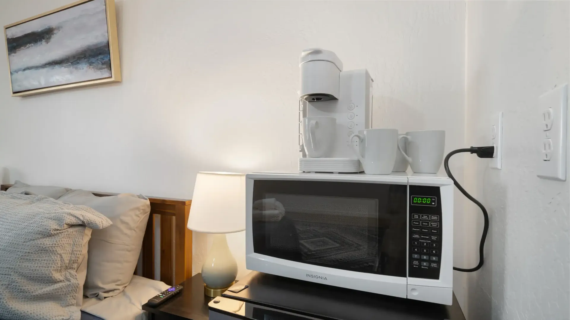 Microwave, coffee maker, and mugs on a nightstand in a room with bed and artwork.