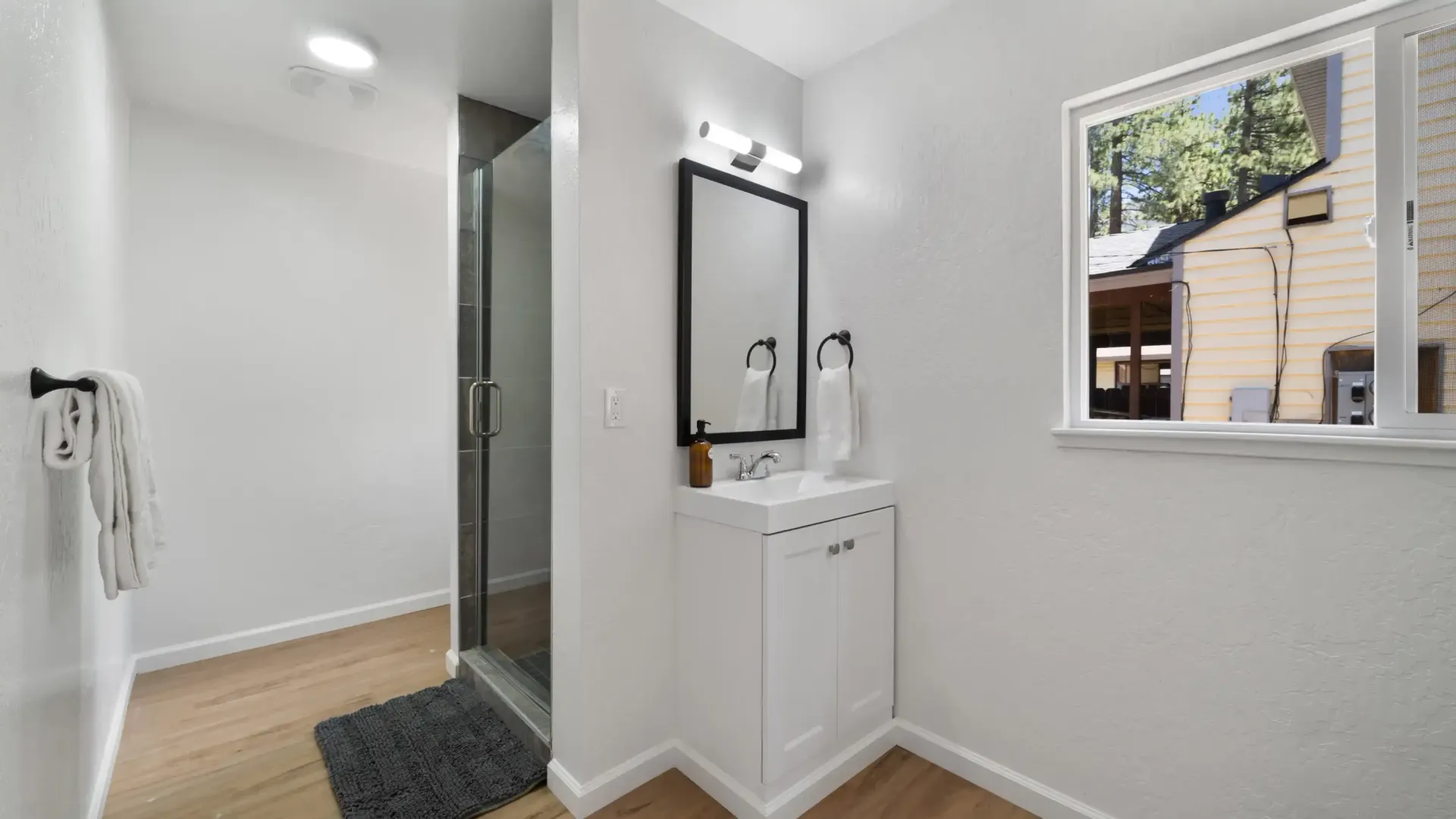 Bathroom with a white vanity, black-framed mirror, and shower. Window provides natural light.