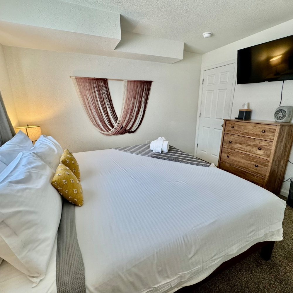 Bedroom with white bed, wooden dresser, and mounted TV on a white wall.