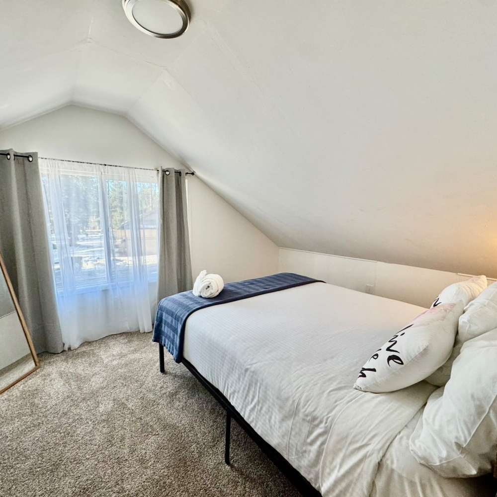 Bedroom with a bed, window, mirror, and sloped ceiling. White bedding and light gray curtains.