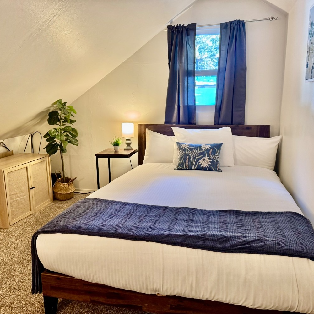Bedroom with a bed, small window with blue curtains, and a side table with a lamp.