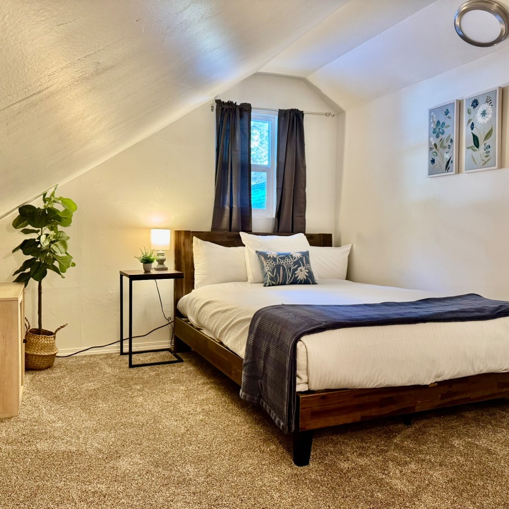 Bedroom with bed, small window, side table, plant, and wall art. Soft, neutral colors.