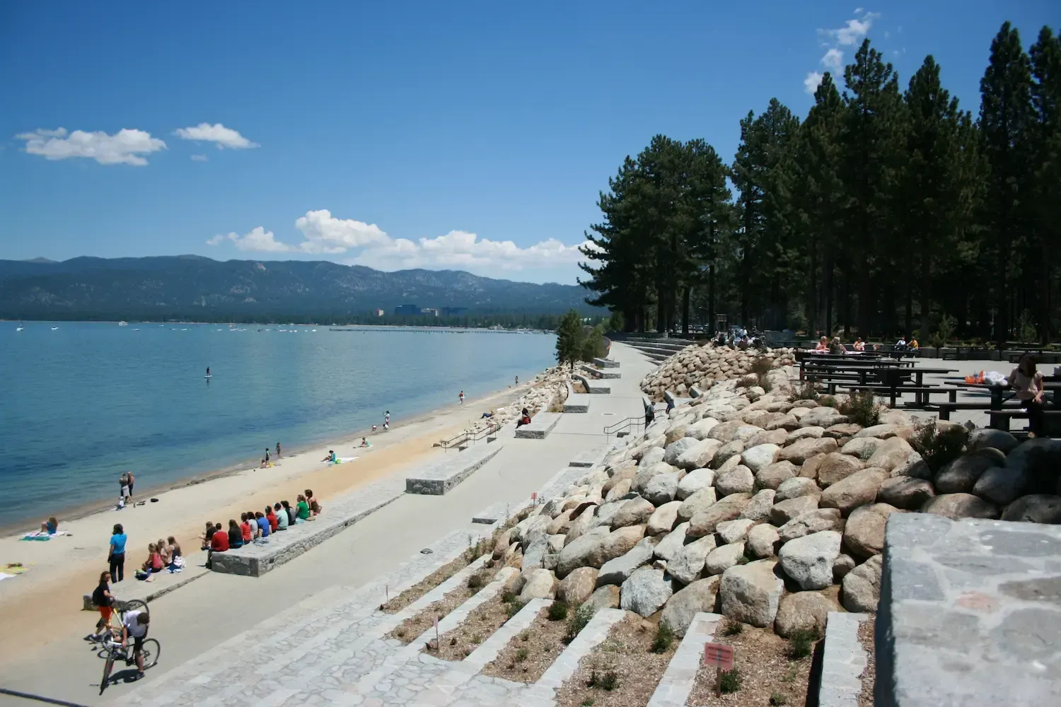 Beach with people, blue water, and forested shoreline on a sunny day.