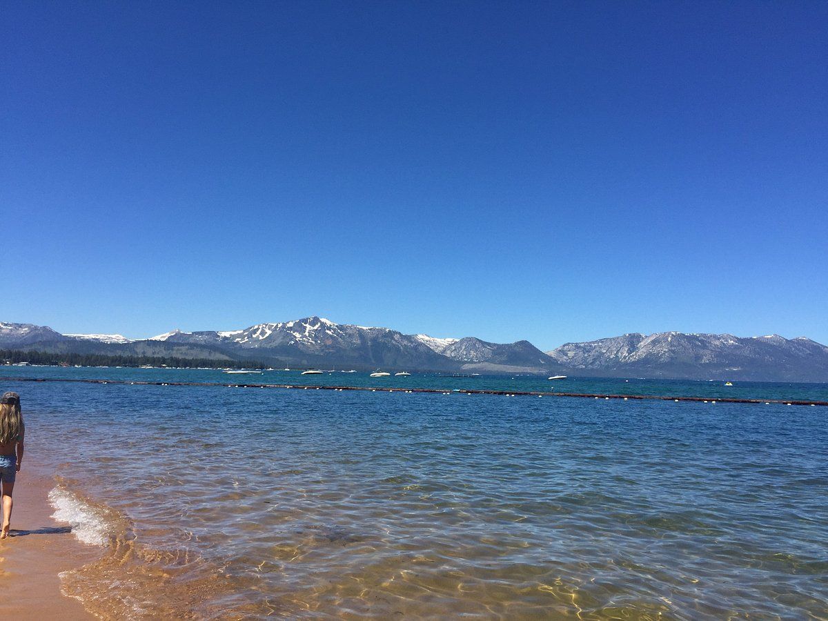 Person walking on a sandy beach; blue water, snow-capped mountains, clear sky.
